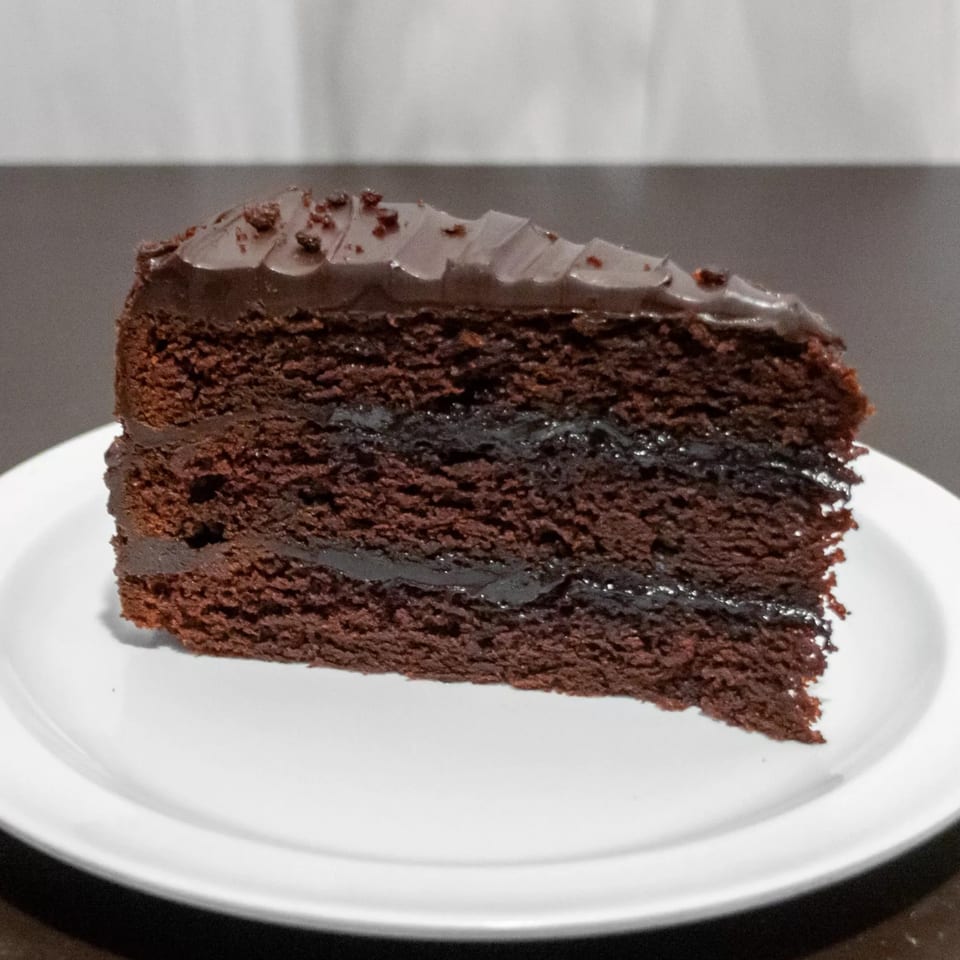 A rich slice of chocolate cake with chocolate frosting and filling on a white plate.