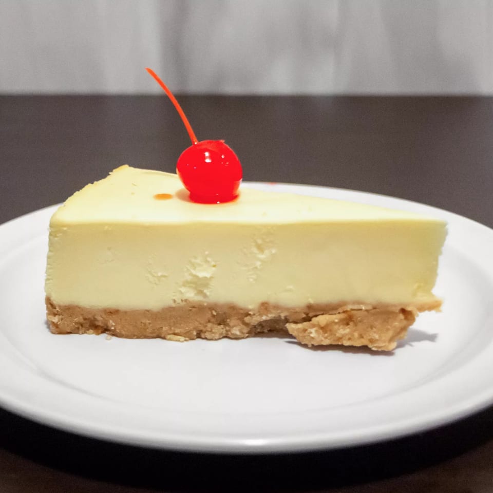 A slice of cheesecake with a graham cracker crust and a red maraschino cherry on top.